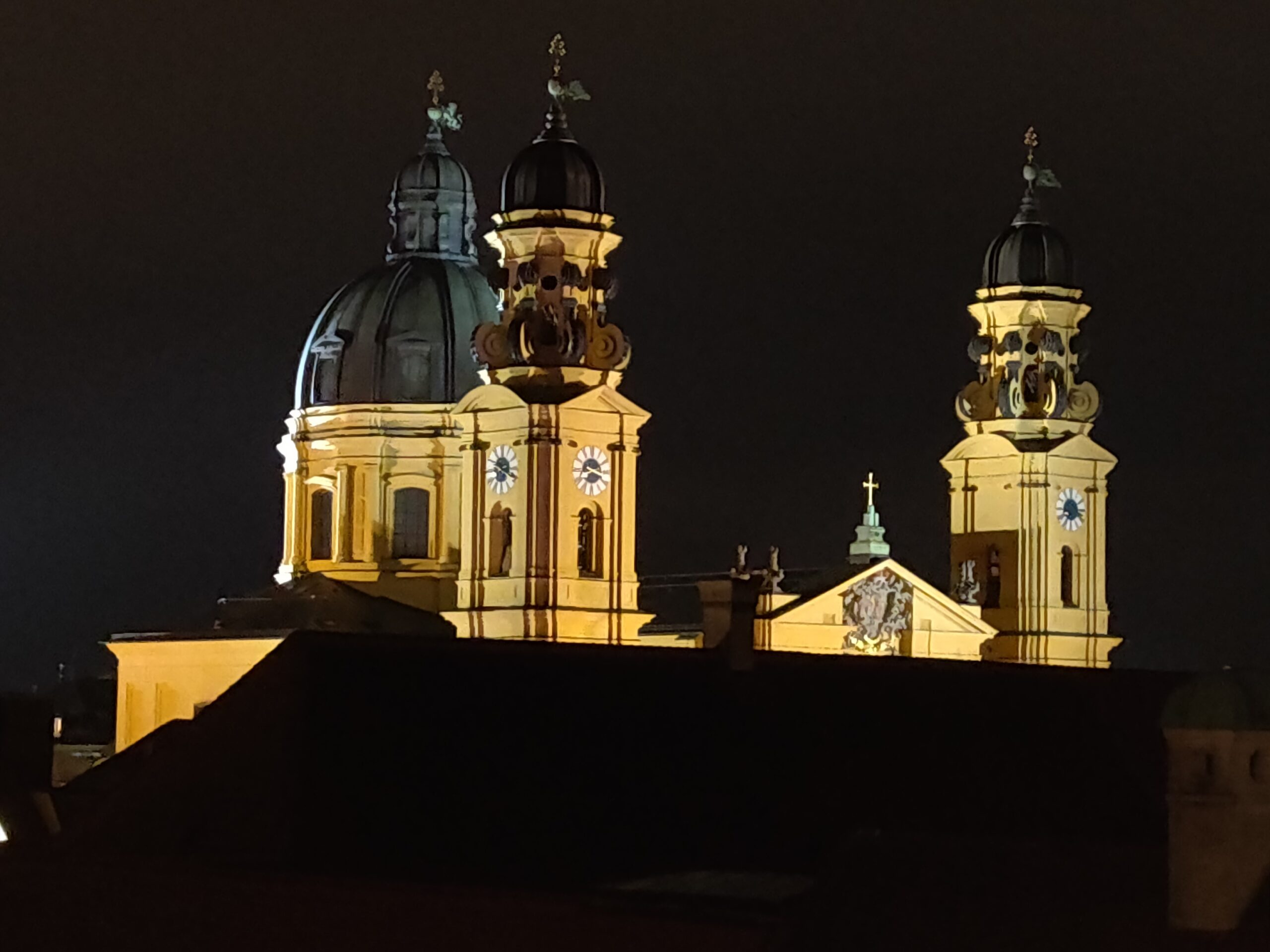 Eine prächtig beleuchtete Barockkirche mit zwei Türmen und einer Kuppel bei Nacht, deren gelbe Fassade dramatisch gegen den dunklen Himmel hervortritt.