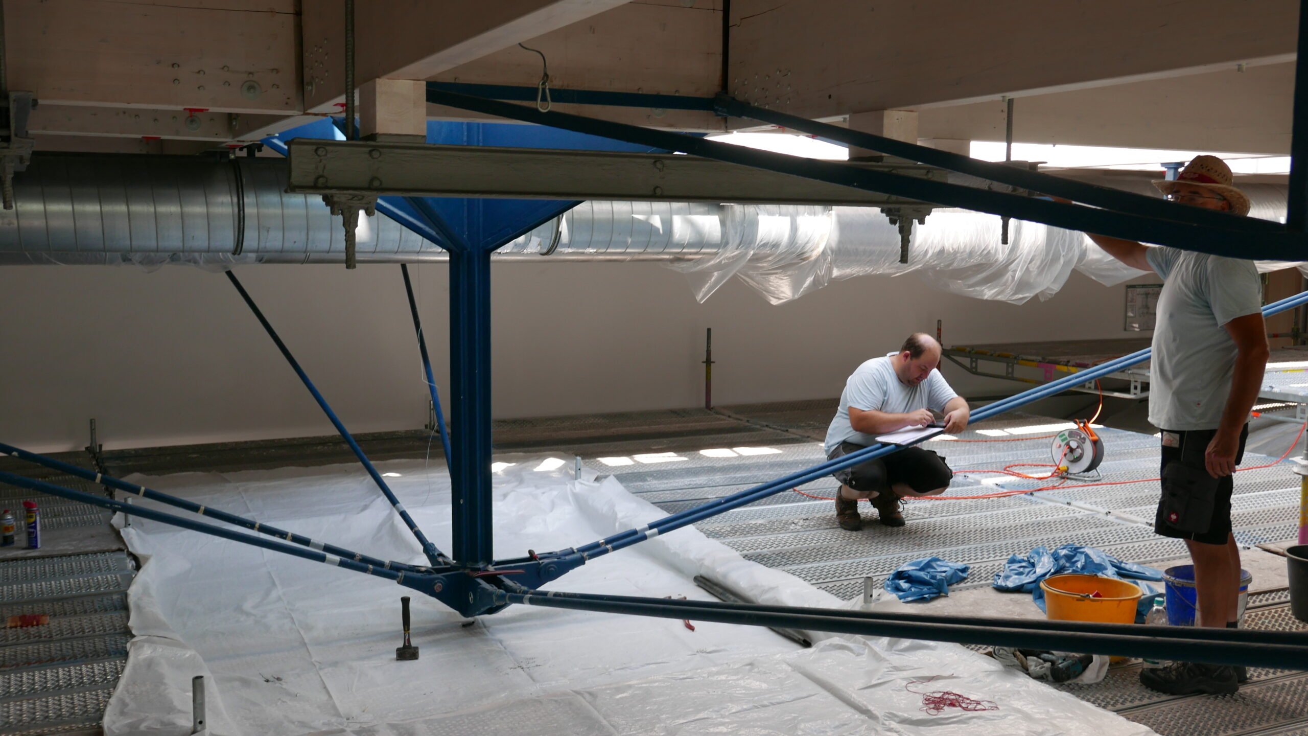 In einer modernen Industriehalle arbeiten zwei Handwerker an der Installation von blauen Rohrleitungen unter einer Stahlkonstruktion.