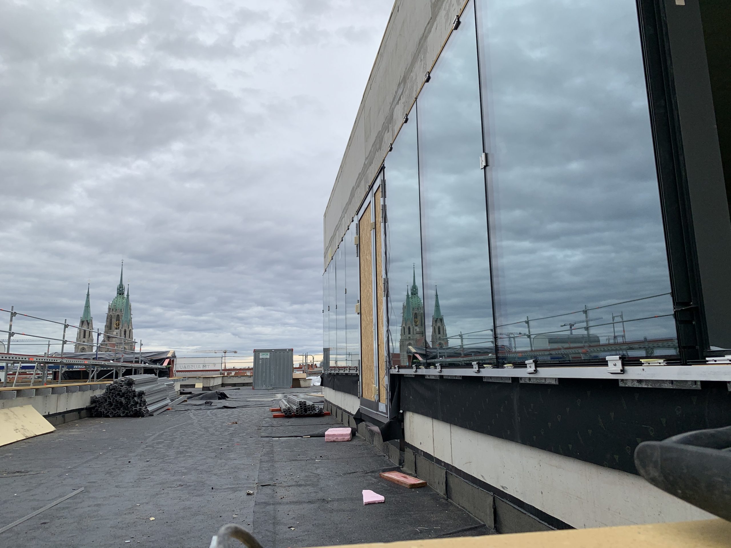 Ein modernes Gebäude mit Glasfassade spiegelt historische Kirchen wider, die sich auf einem urbanen Dach erheben.