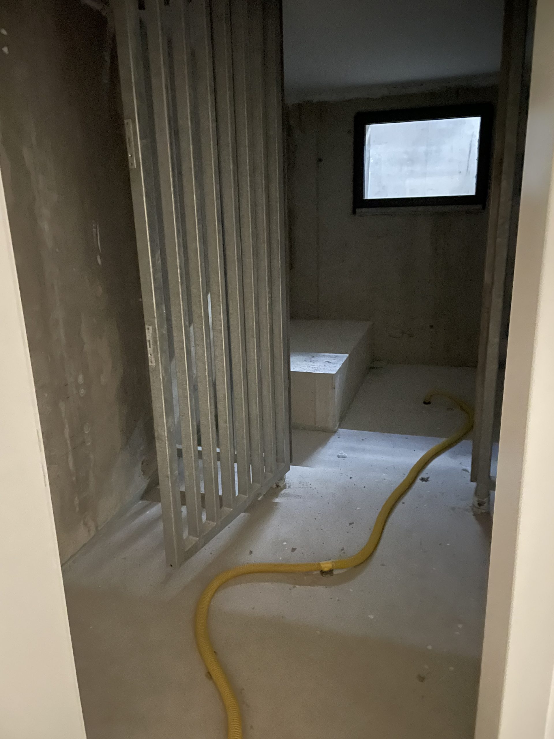 Ein unfertiger Innenraum mit Betonwänden, Fenster, Stahlrahmen und gelber Schlauch auf dem Boden.