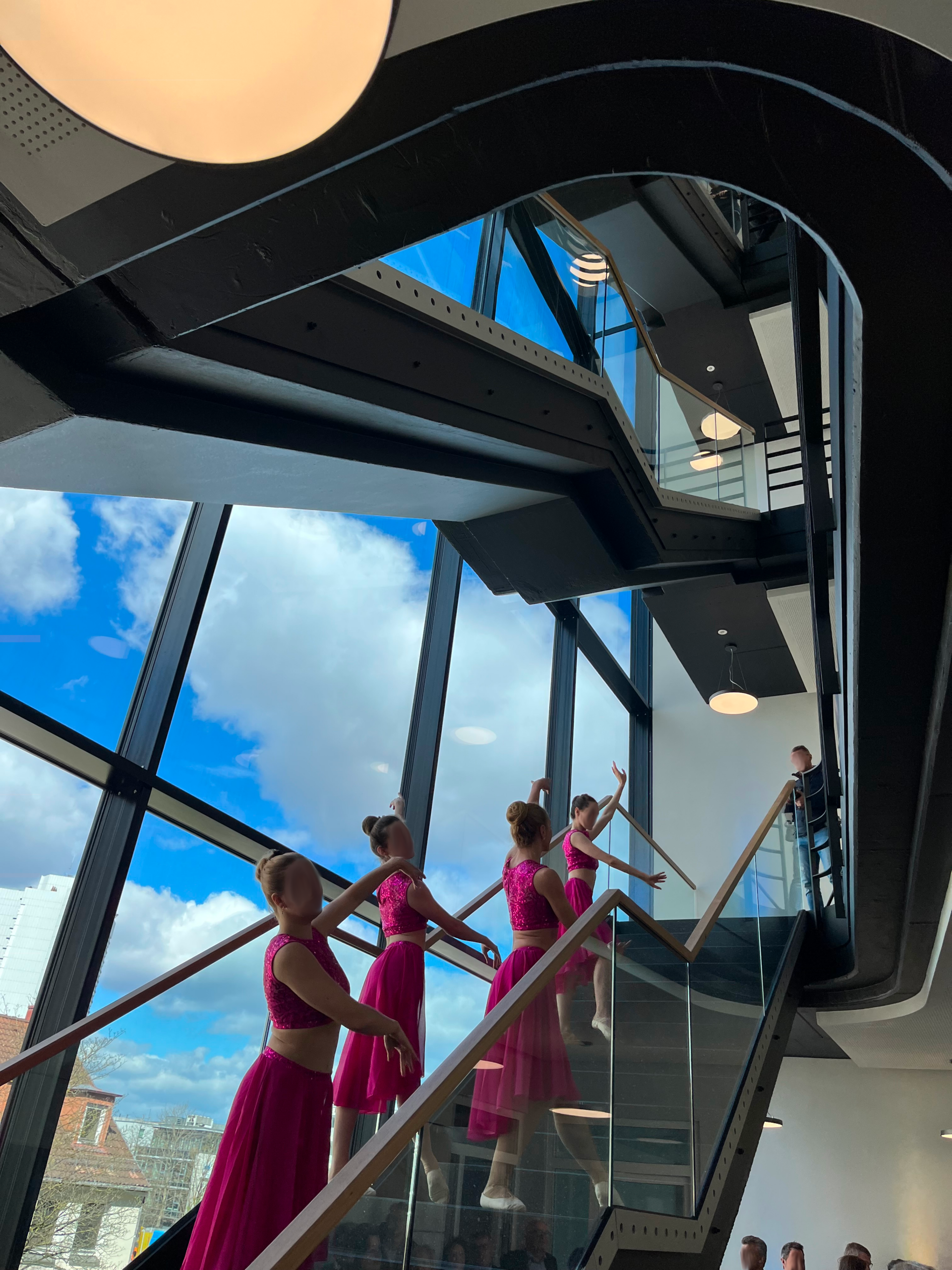 Junge Tänzerinnen in pinken Outfits posieren auf einer modernen Treppe vor großen Glasfassaden mit Blick auf den blauen Himmel.