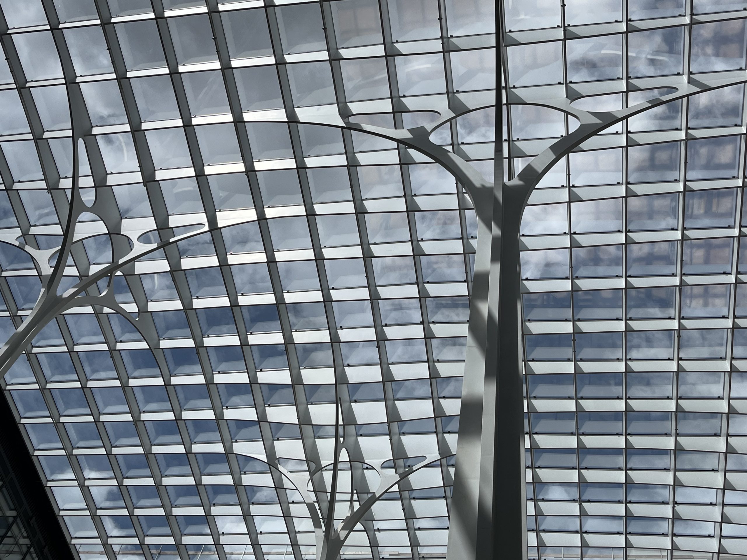 Eine moderne Glasfassade mit geometrischem Rasterwerk bildet das Dach eines zeitgenössischen Gebäudes, durchbrochen von stilisierten metallenen Baum-Skulpturen.