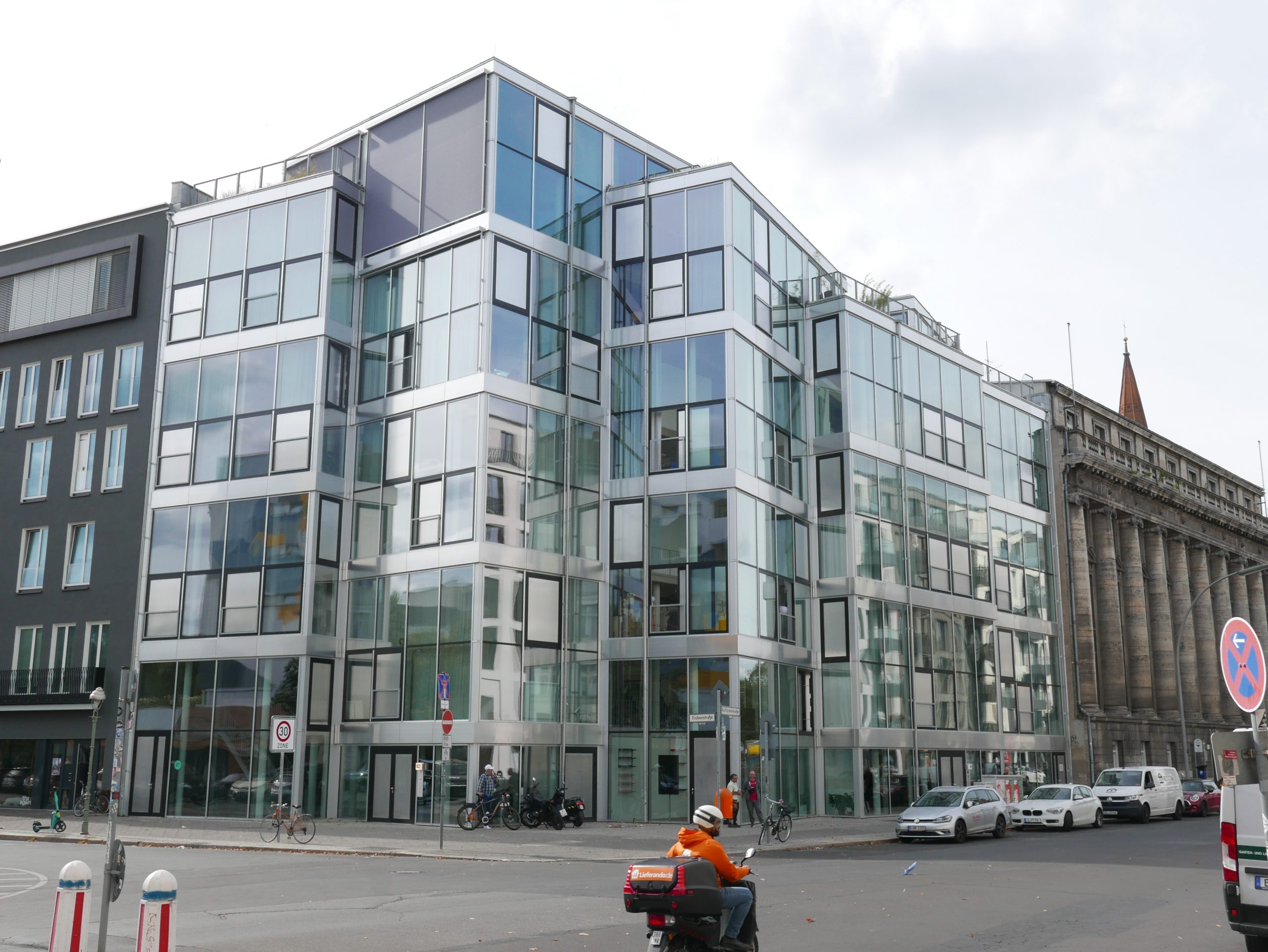 Ein modernes Glasgebäude mit weißem Rahmen und großflächigen Fenstern steht in einer urbanen Umgebung neben historischen Backsteinbauten.