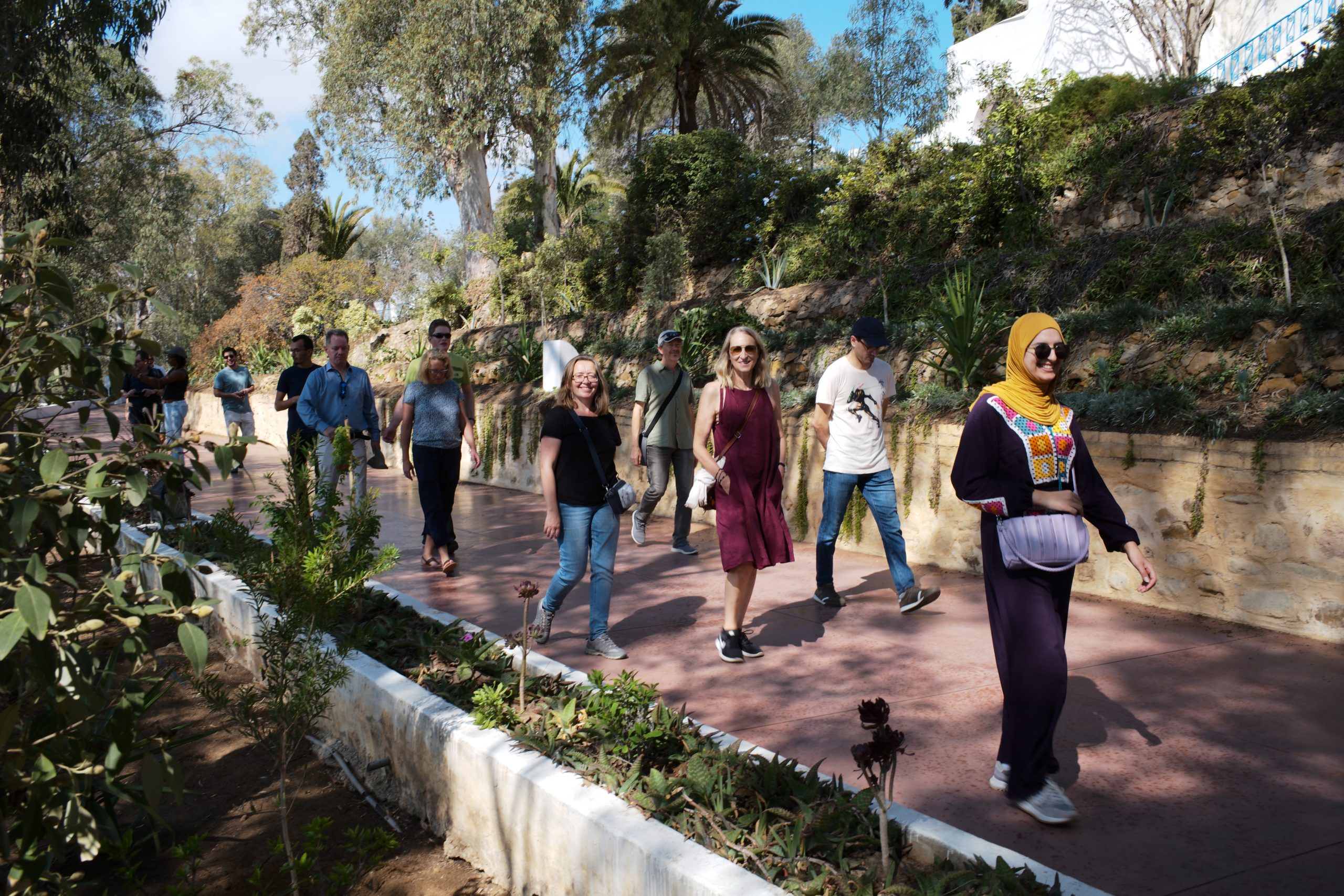 Eine Gruppe von Touristen spaziert durch einen terrassierten Garten mit Palmen und Pflanzen neben einer historischen Steinmauer.