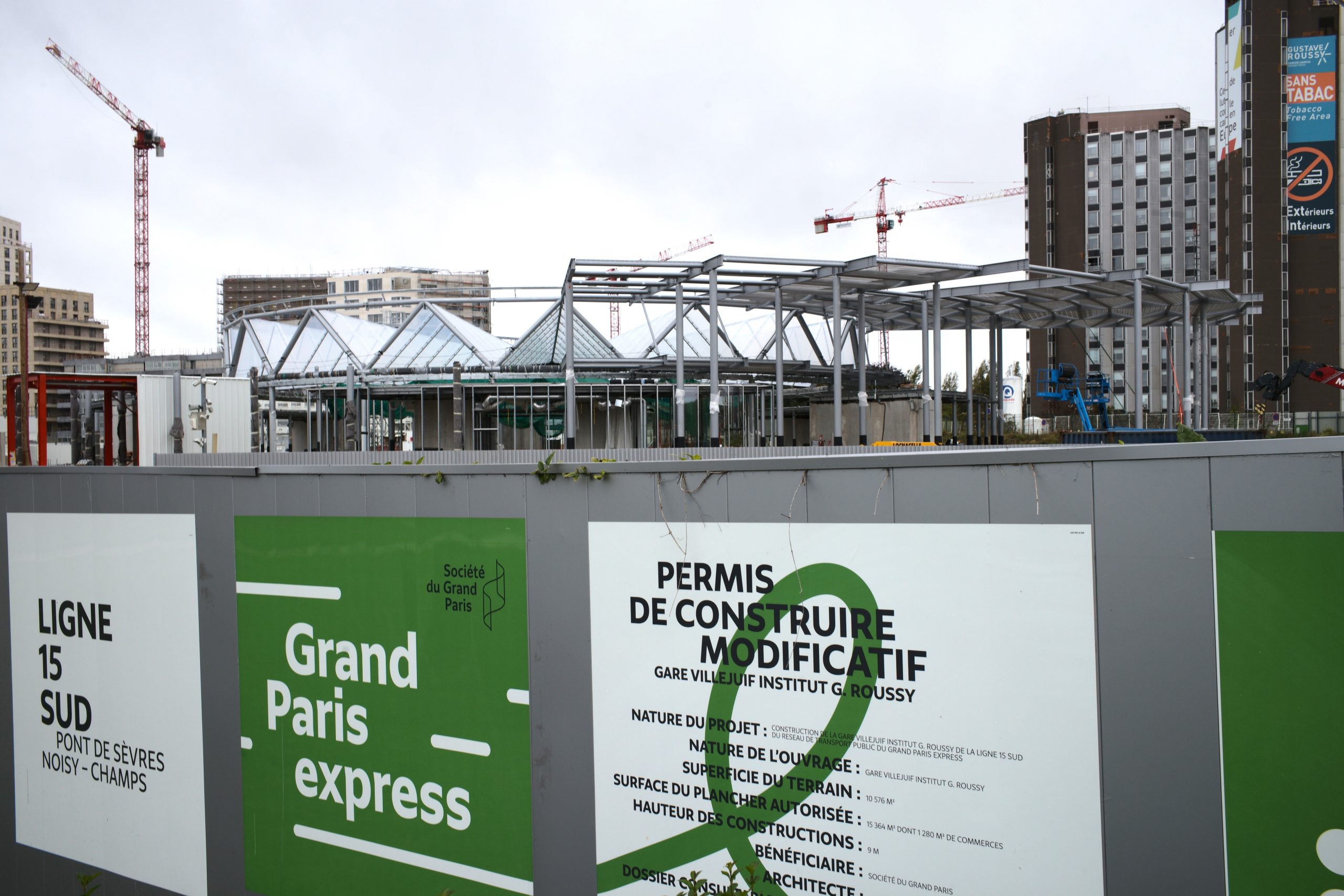 Eine moderne Baustelle in Paris mit Stahlkonstruktion und Glasdach, umgeben von grauen Bauschutzzäunen und Kränen.