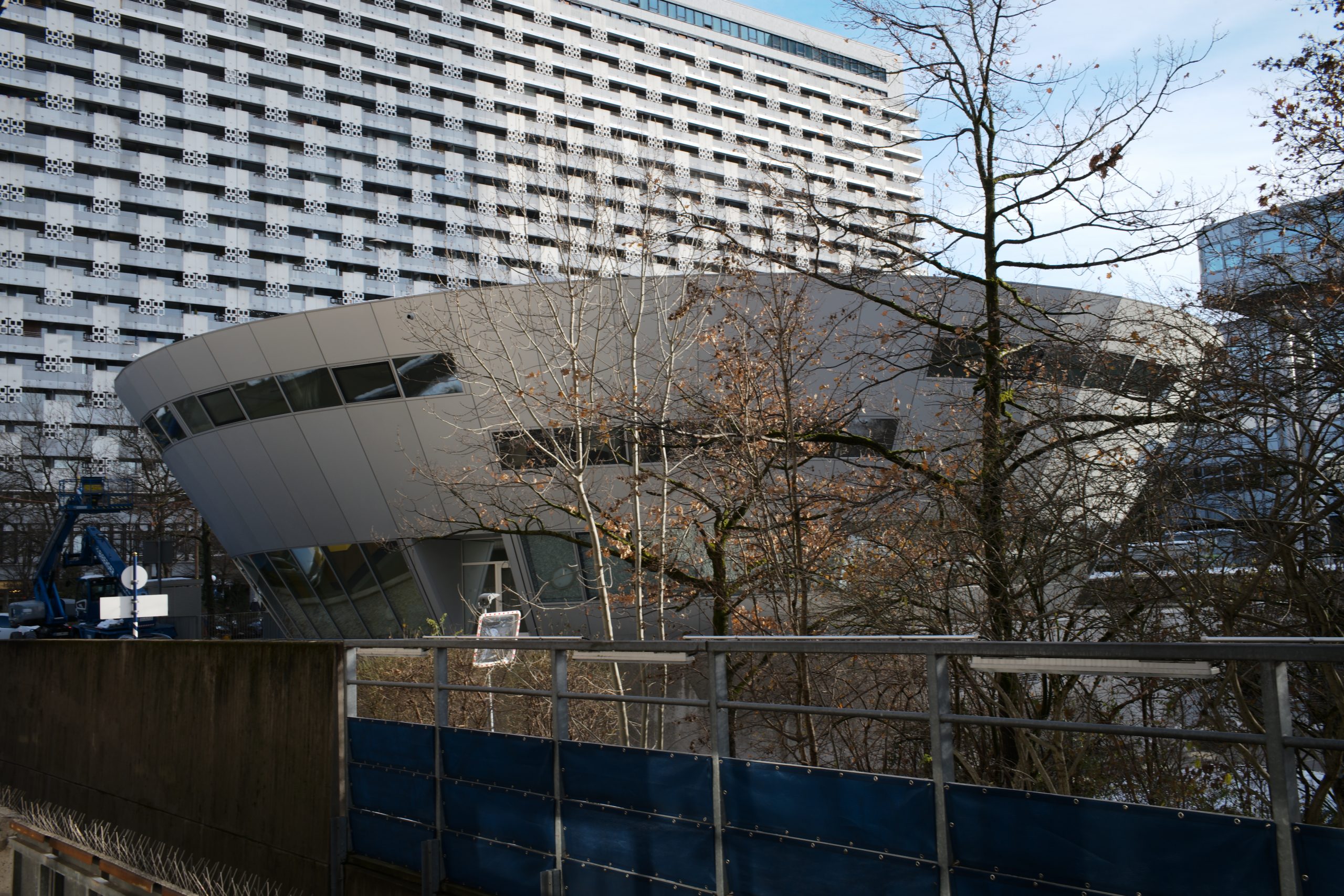 Ein modernes, futuristisches Gebäude mit gekrümmter weißer Fassade steht vor einem großen Wohnblock mit charakteristischem Rastermusters.