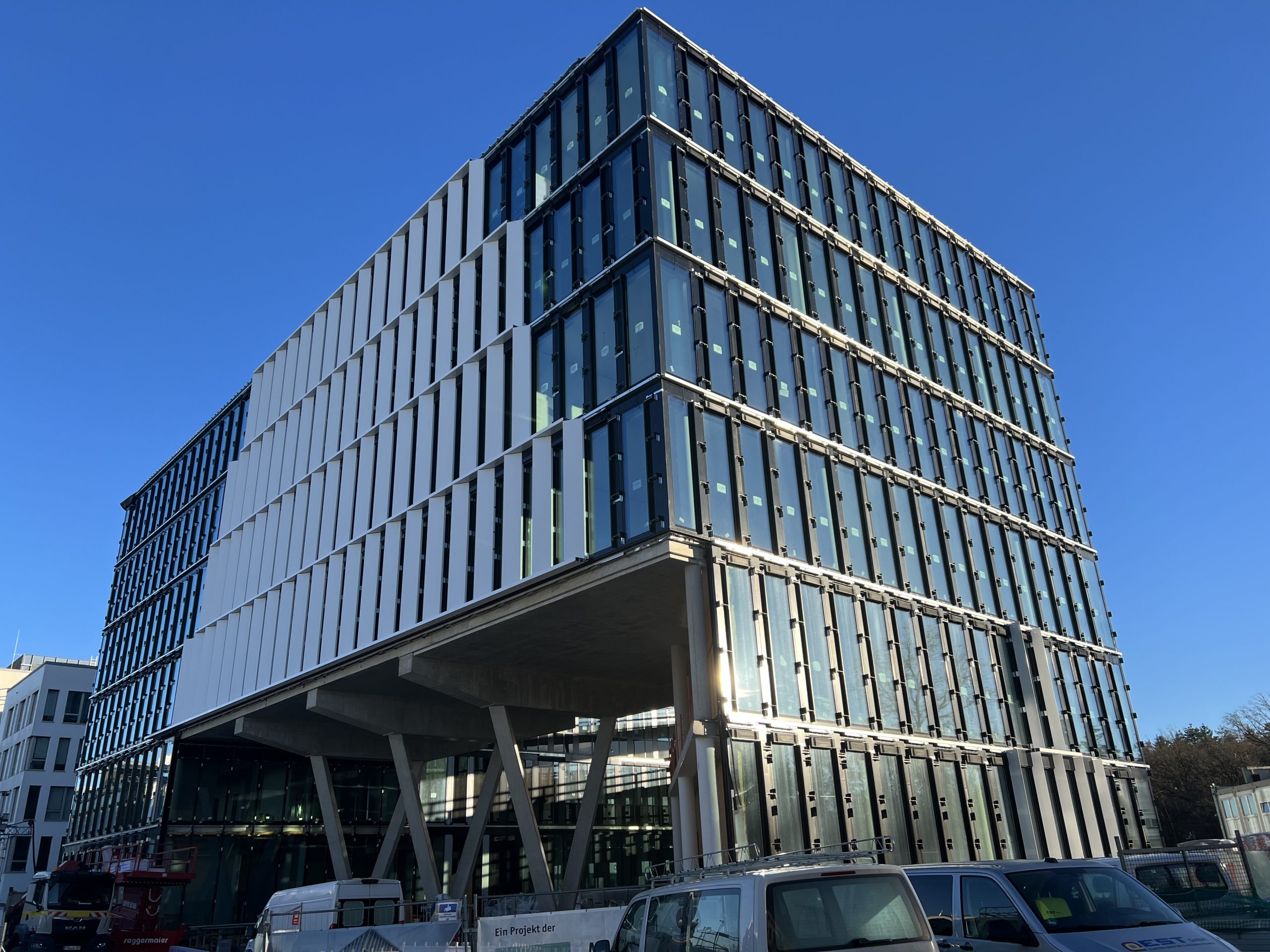 Ein modernes Hochhaus mit markanter Gittersichtfassade aus grauem Metall und Glas, das auf massiven Betonstützen ruht und unter klarem blauen Himmel fotogen wirkt.