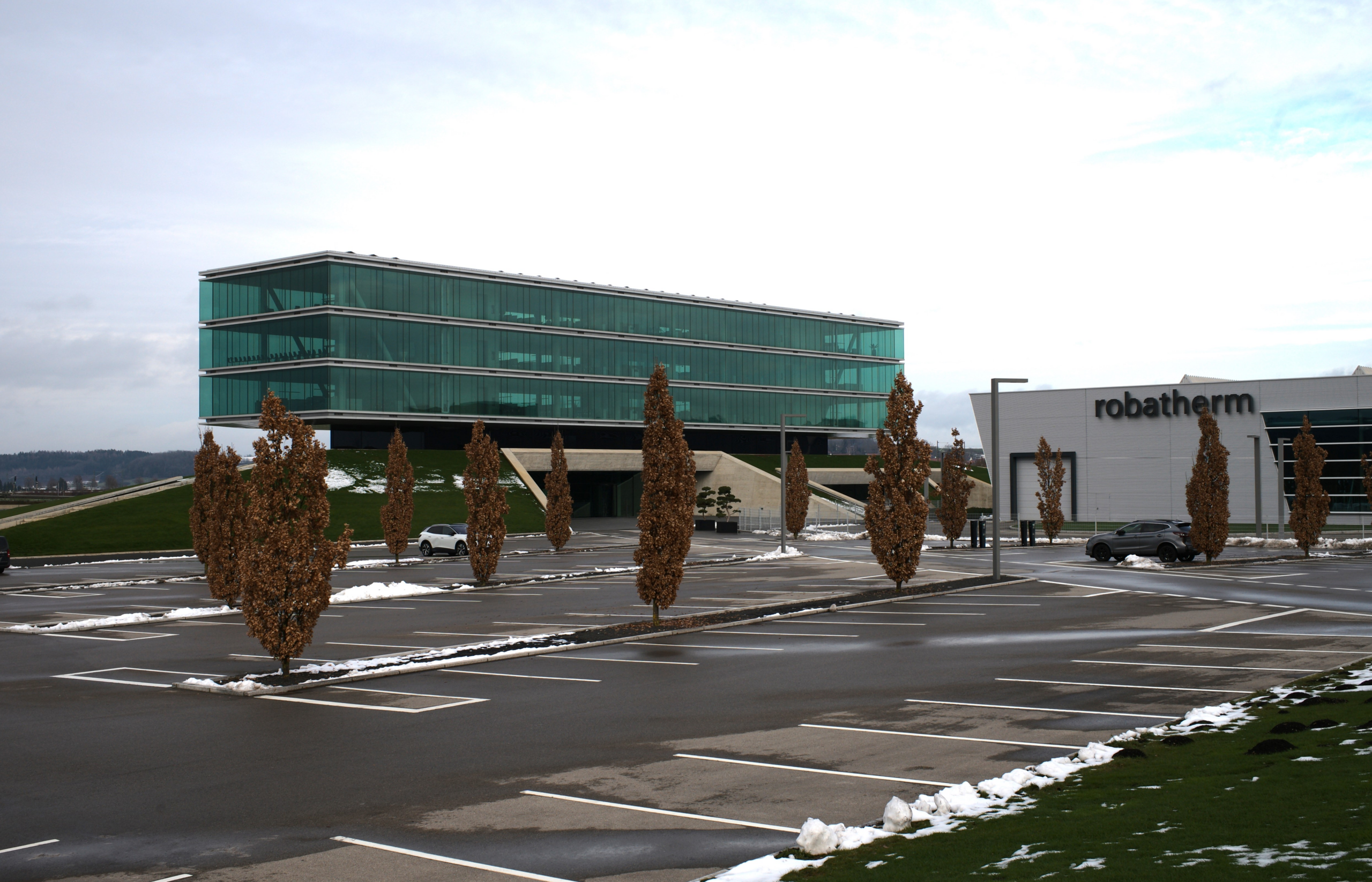 Ein modernes Gebäude mit grüner Glasfassade und horizontalen Bändern steht auf Stelzen neben einem weißen Robatherm-Gebäude in einer Parkanlage.