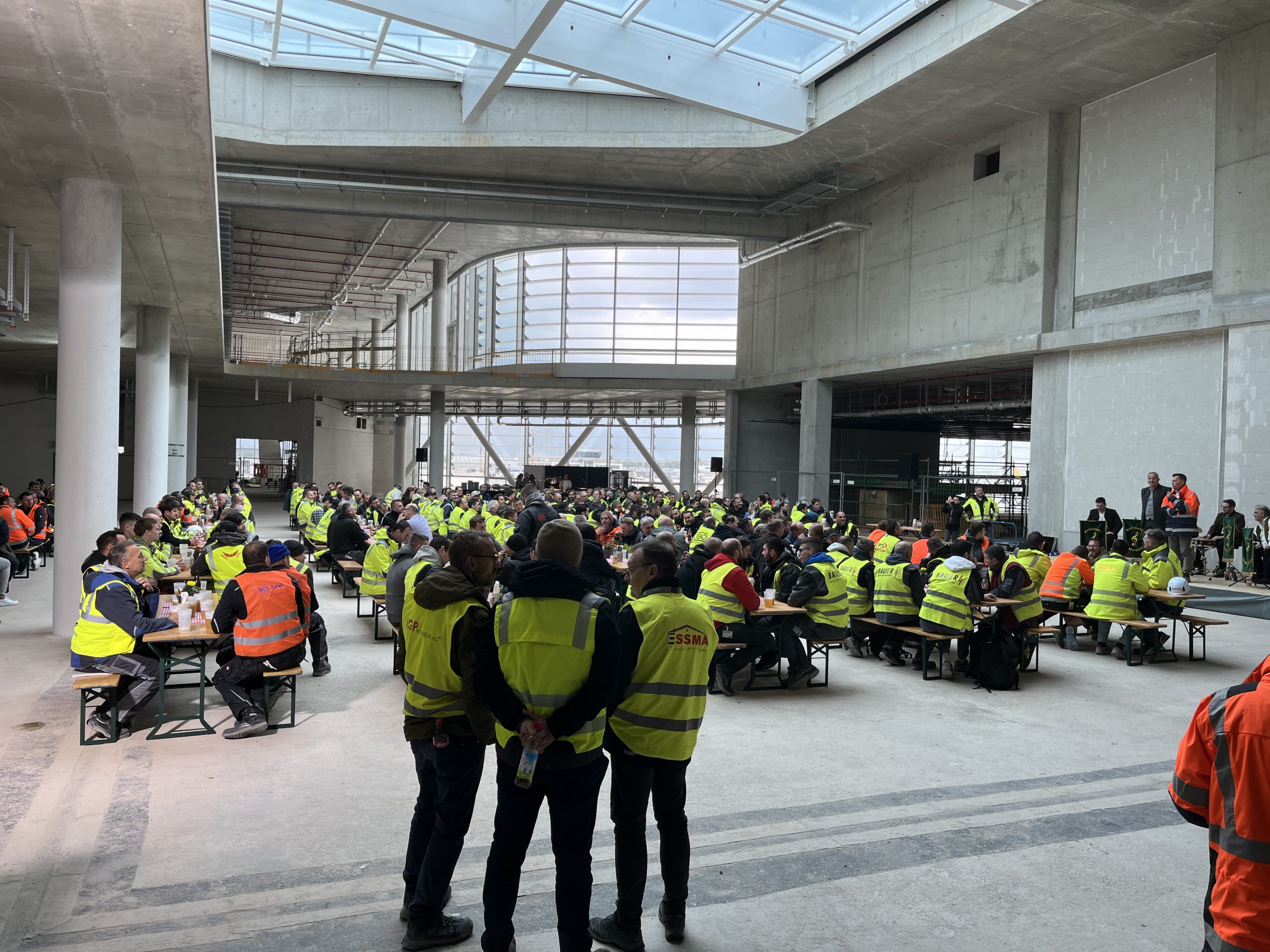 Ein modernes Betongebäude mit großen Glasfenstern und Oberlichtern beherbergt eine Versammlung von Personen in Warnwesten.