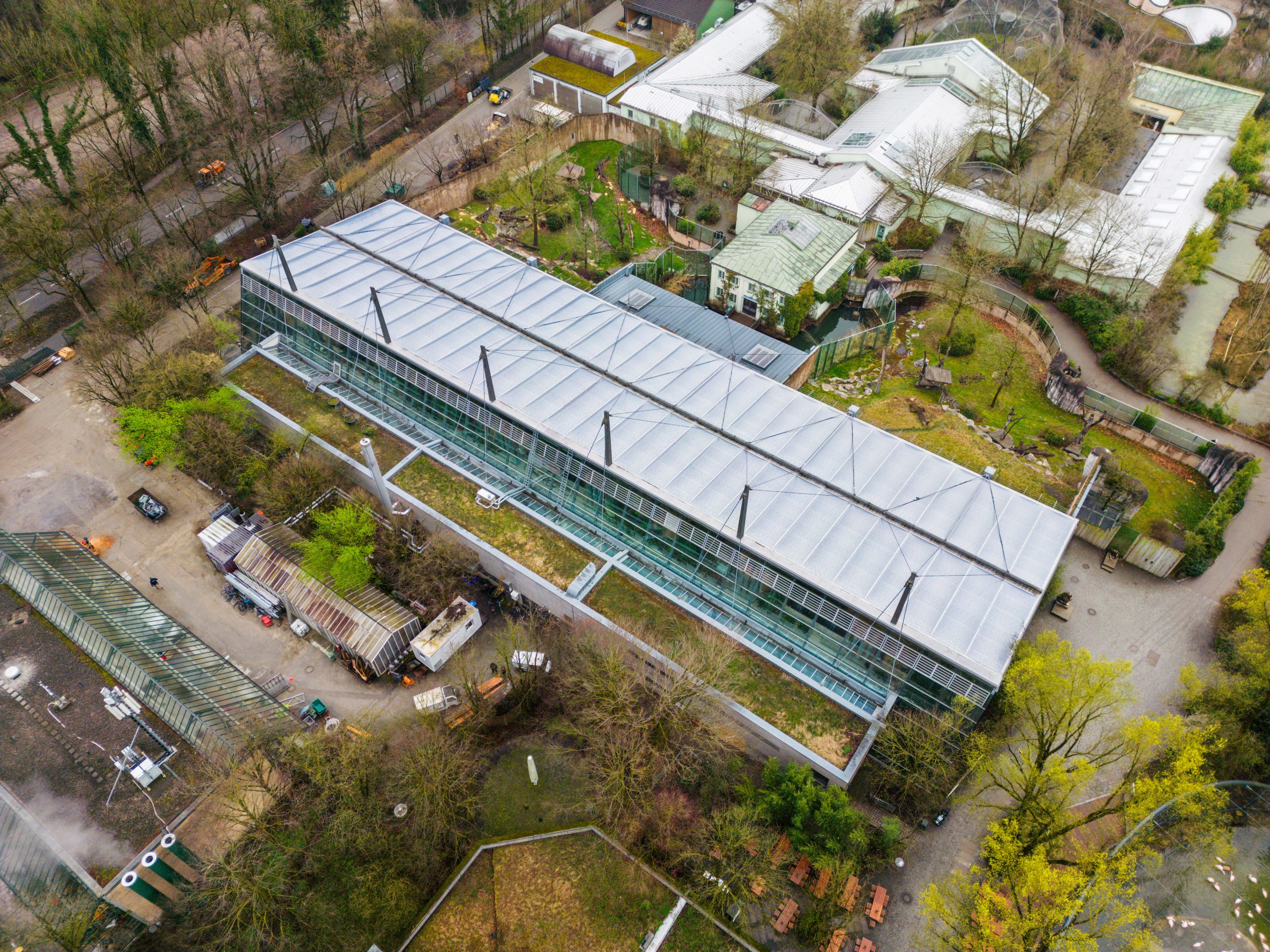 Ein modernes Gewächshaus mit weißem Membrandach und grüner Glasfassade liegt eingebettet in eine grüne, waldige Umgebung mit umgebenden Gartenbaubetrieben.