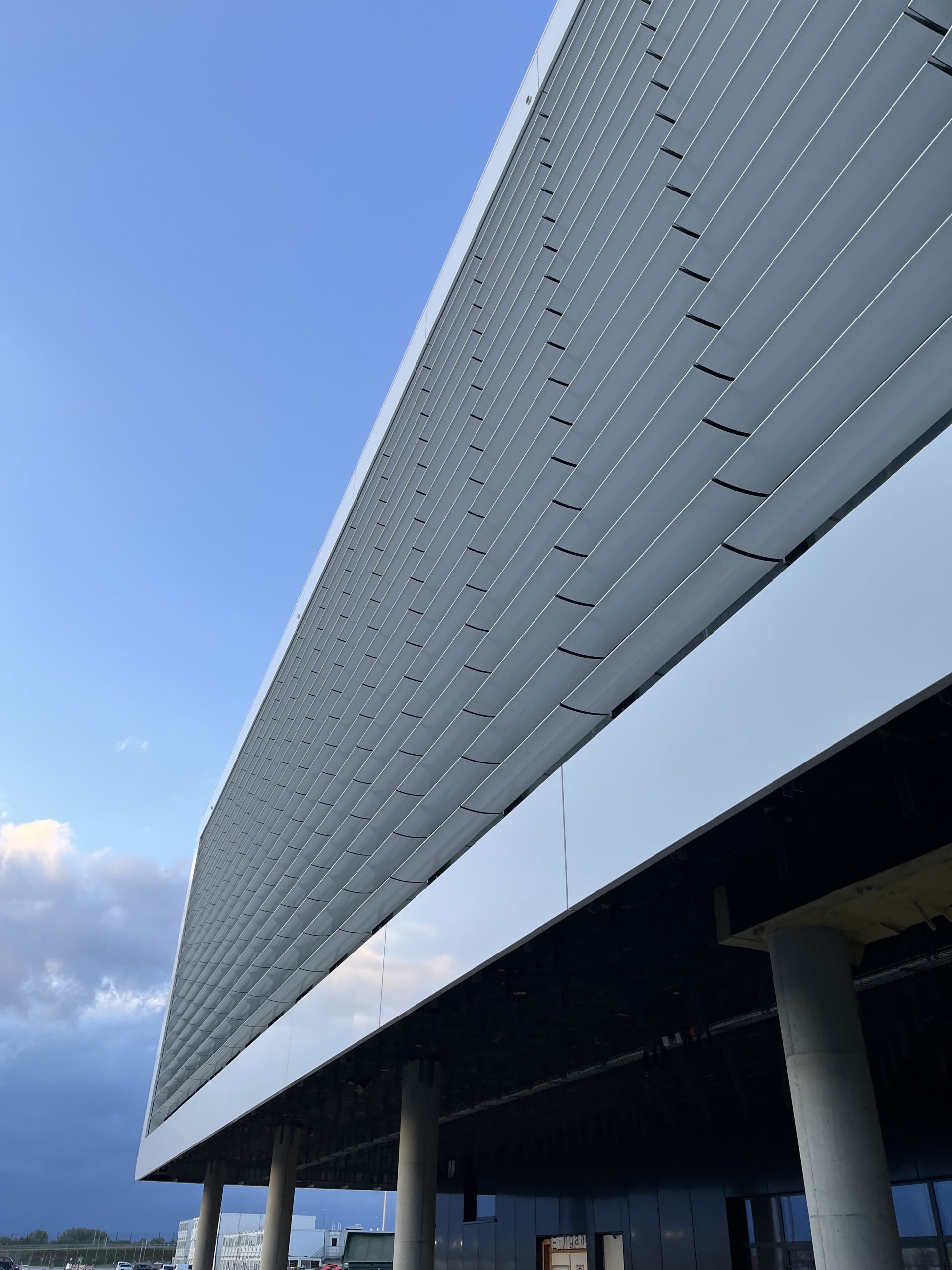 Ein modernes Gebäude mit charakteristischer grauer Metallverkleidung schwebt auf Betonstützen über einem dunklen Untergeschoss unter blauem Himmel.