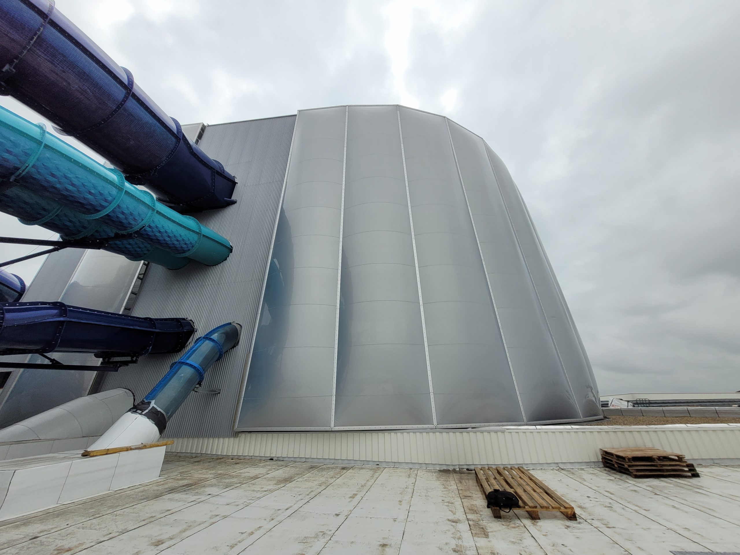 Ein großer, zylindrischer Industriebehälter aus Metall mit vertikalen Verstrebungen steht auf einem Flachdach neben blauen Rohren.