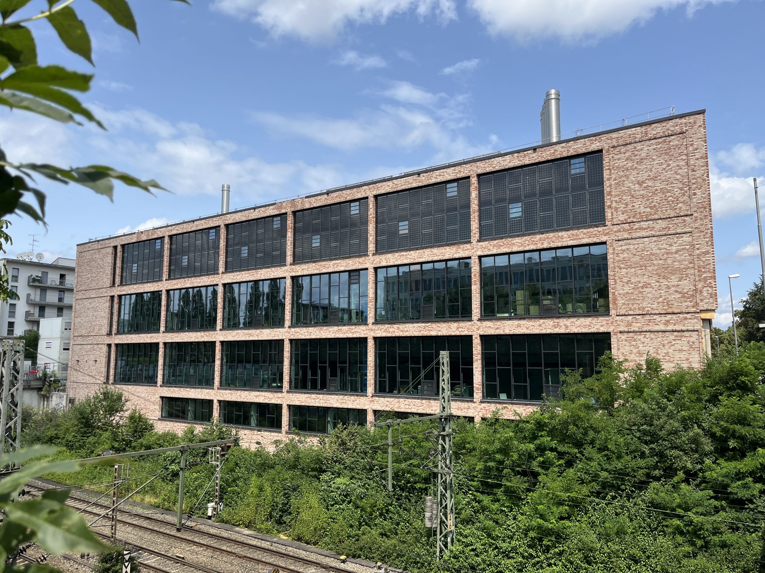 Ein modernes Backsteingebäude mit großflächigen Fenstern steht neben Eisenbahngleisen, umgeben von grüner Vegetation.