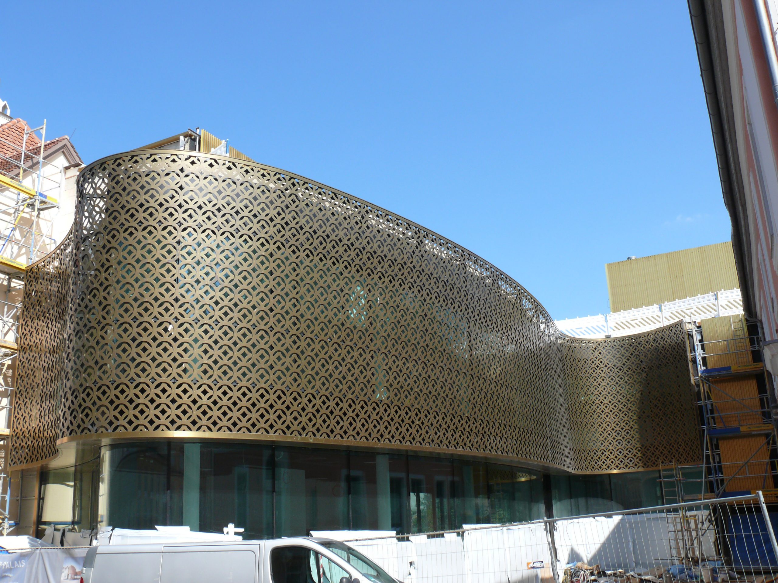 Ein modernes Gebäude mit einer auffälligen geometrischen Metallfassade in Gold- und Brauntönen ist unter klarem Himmel im Bau.