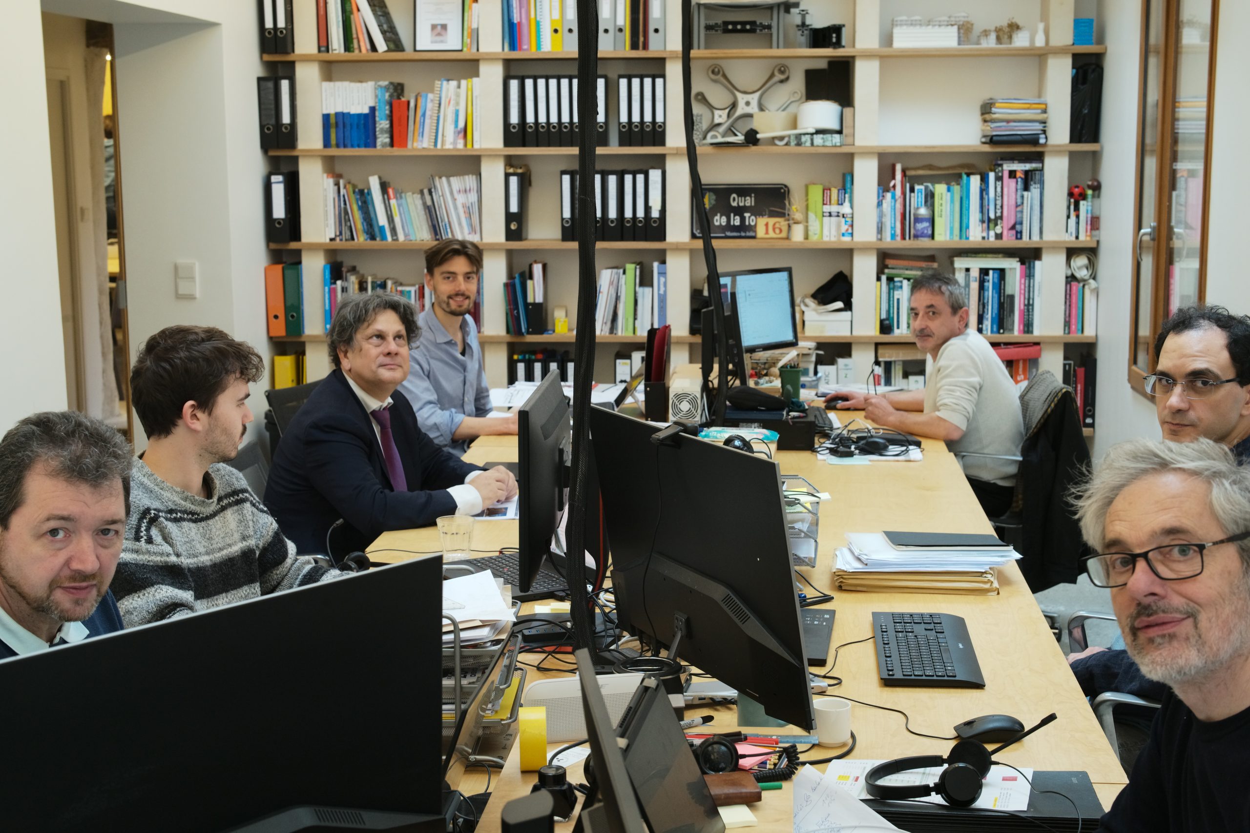 Ein modernes Büro mit hellen Holzregalen, weißen Wänden und zeitgenössischem Design, in dem mehrere Mitarbeiter an Computern arbeiten.