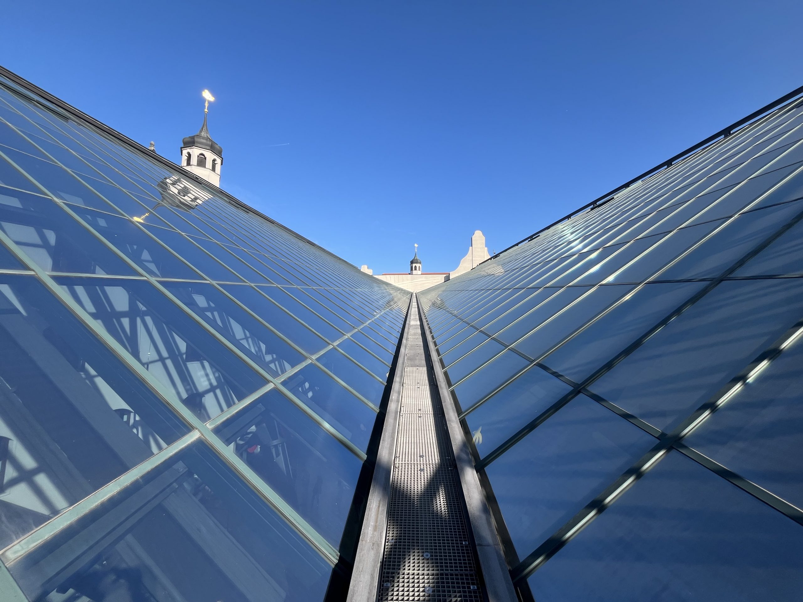 Eine moderne Glasssfassade mit geometrischen Mustern rahmt den Blick auf einen klassischen Kirchturm mit goldenem Kreuz im Hintergrund.