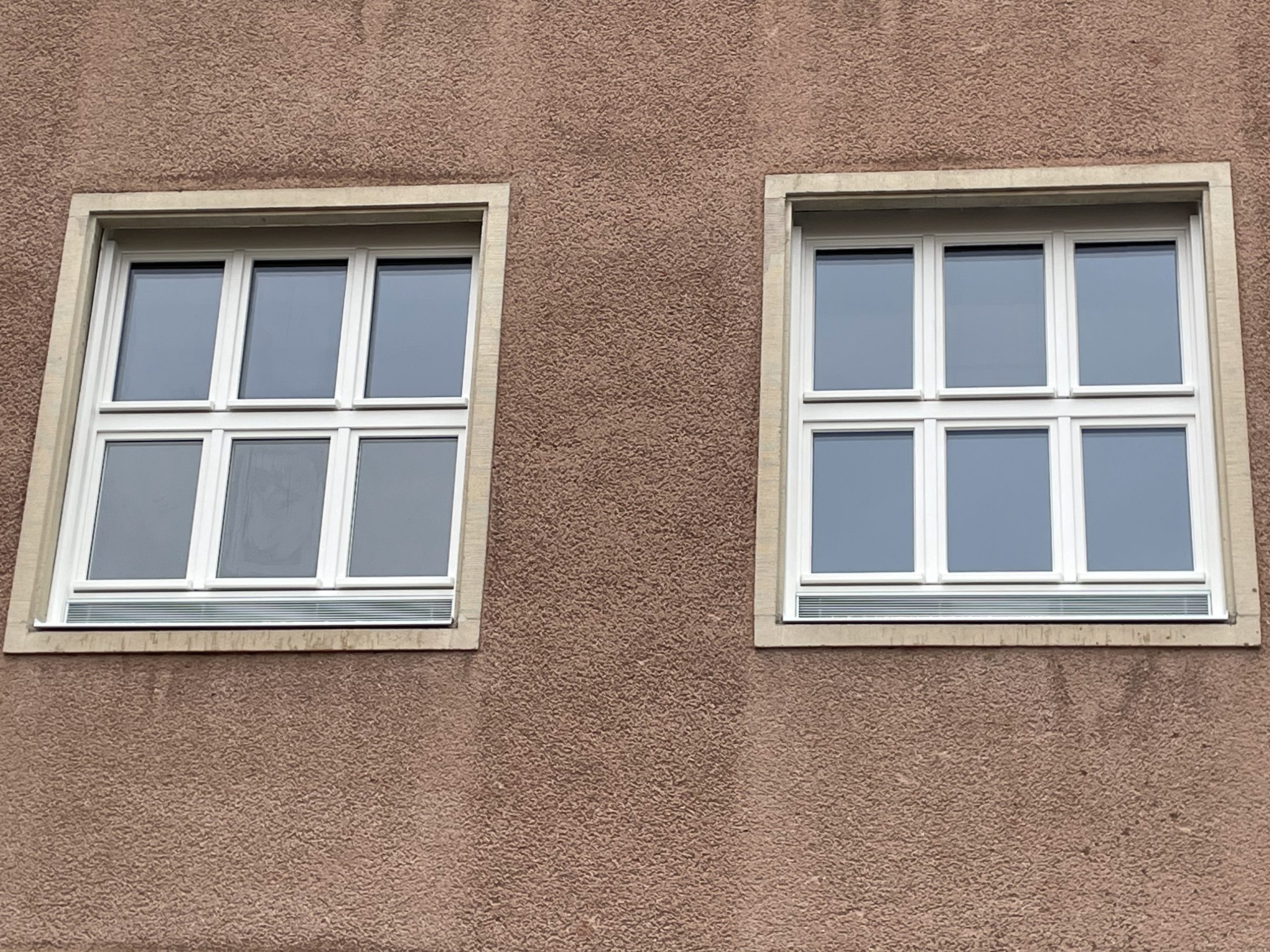 Zwei symmetrische, weiß gerahmte Fenster mit je sechs Scheiben in einer rauen, beigen Putzfassade.