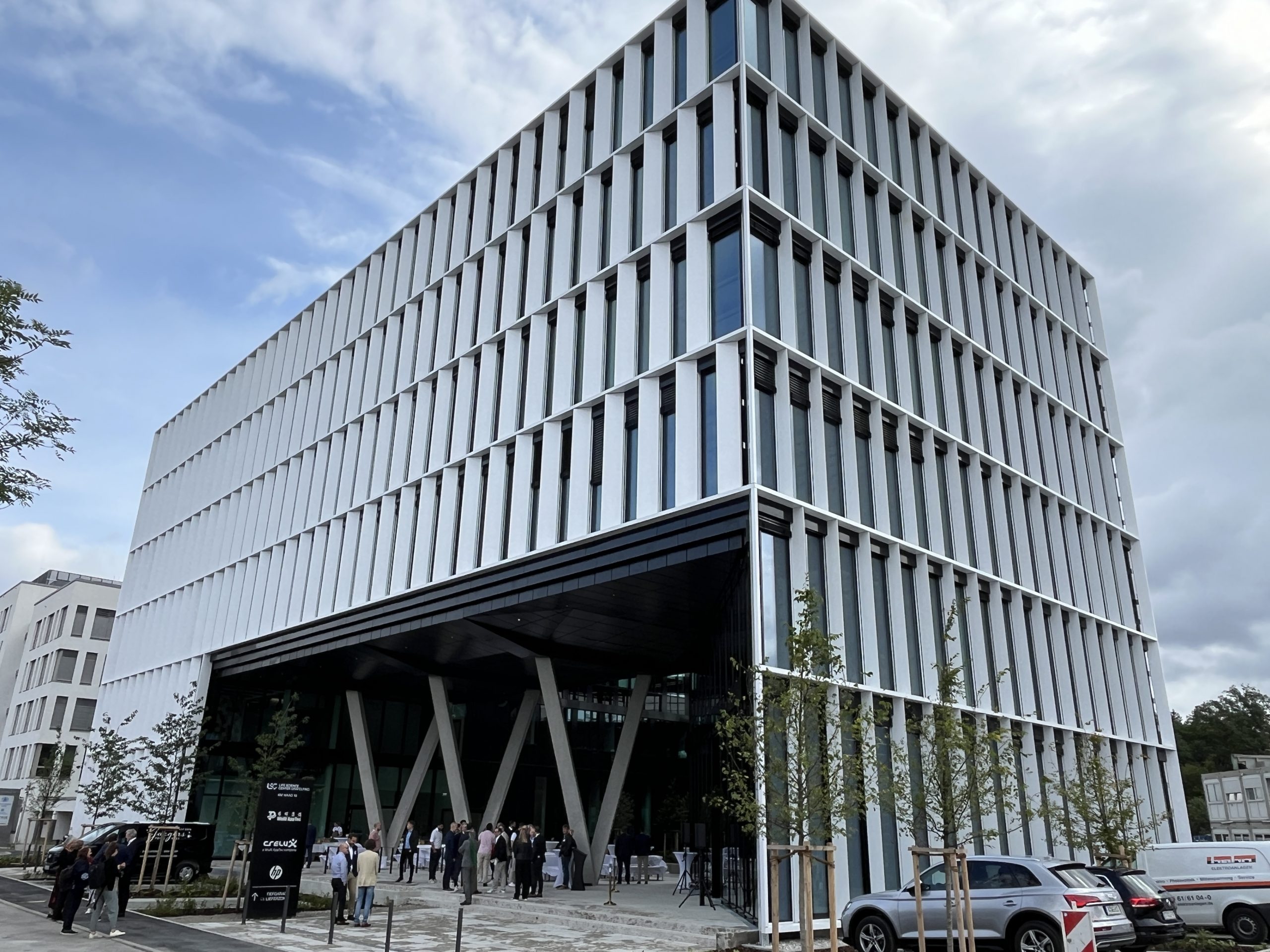 Ein modernes Hochhaus mit vertikaler weißer Lamellenfassade und großflächiger Glasfront im Erdgeschoss, umgeben von Fussgängern und Fahrzeugen.