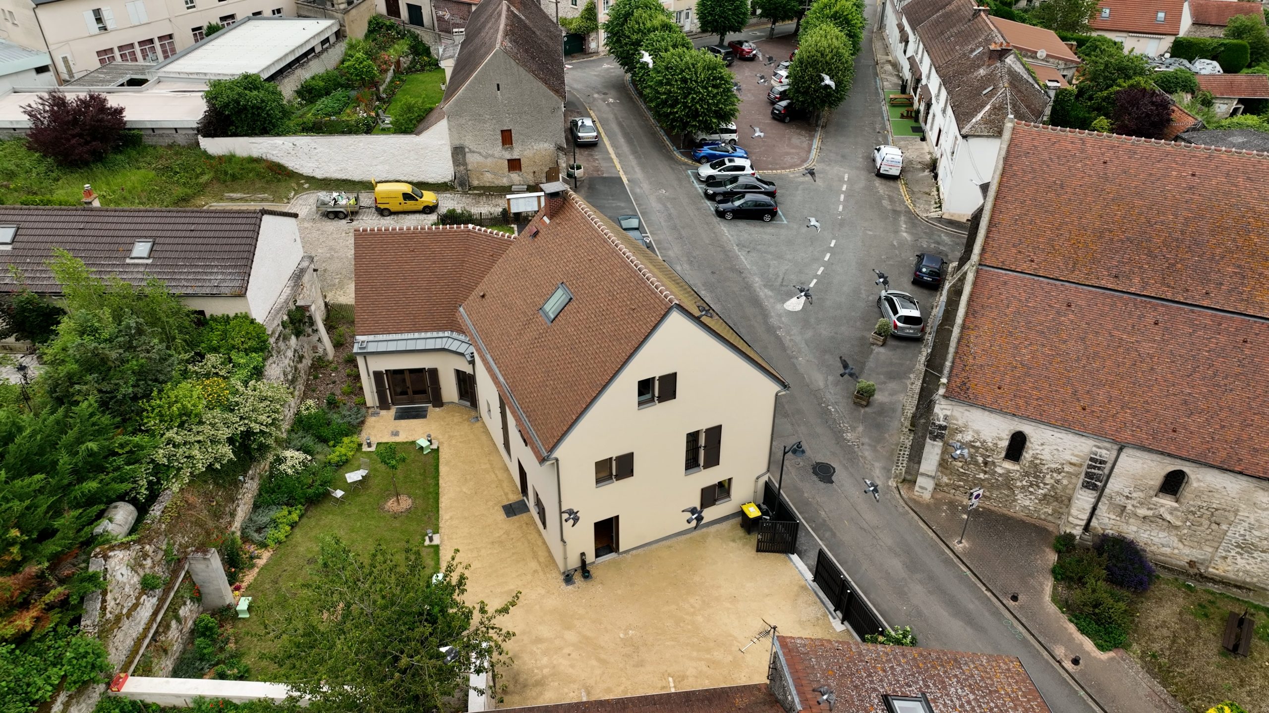 Ein modernes, cremefarbenes Wohnhaus mit Satteldach und großem Sandsteinhof liegt zwischen historischen Gebäuden in einem europäischen Dorf.