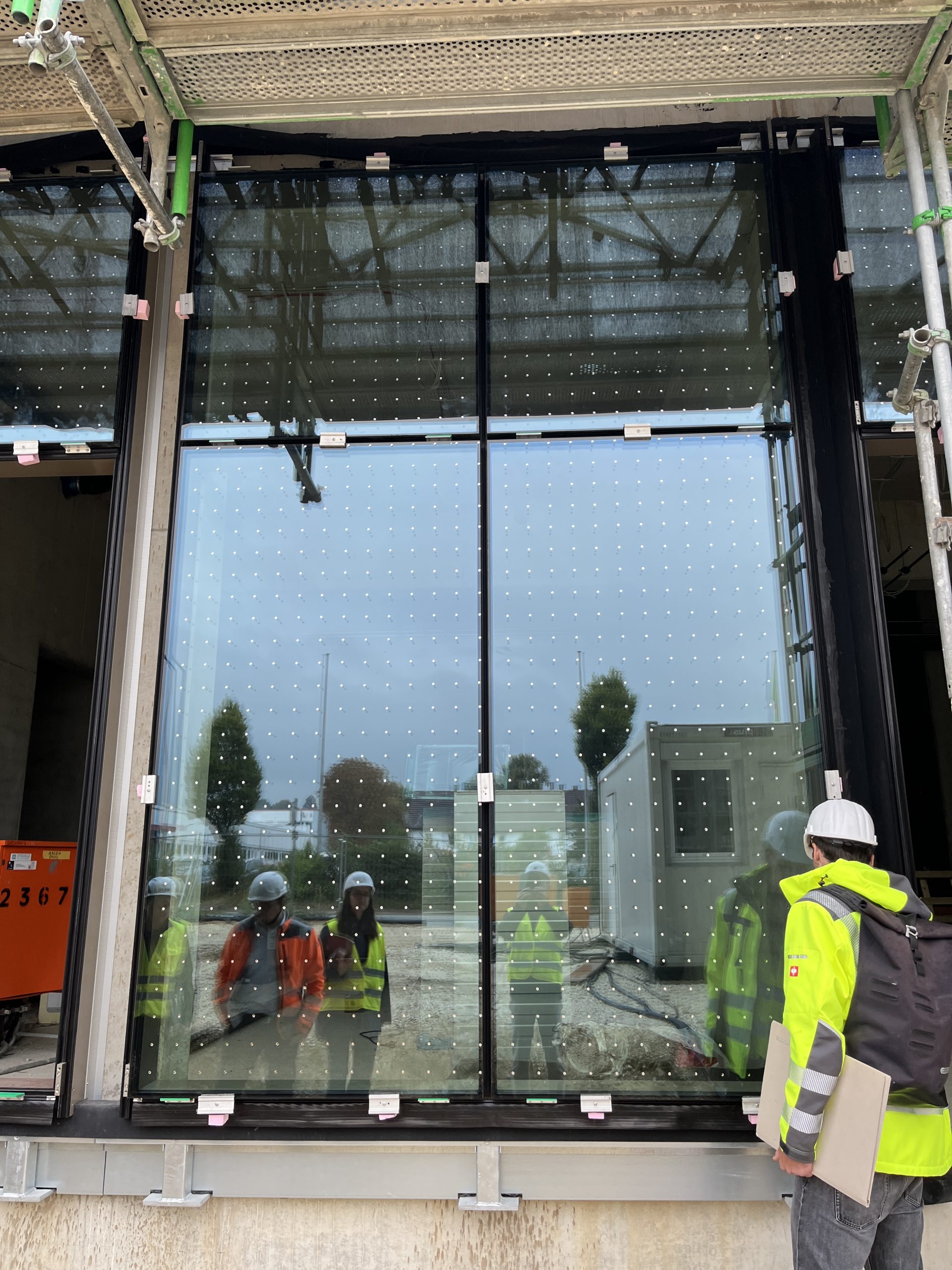 Ein Arbeiter in Schutzausrüstung inspiziert eine großflächige moderne Glasfassade mit Lochblechelementen an einem Gebäude.
