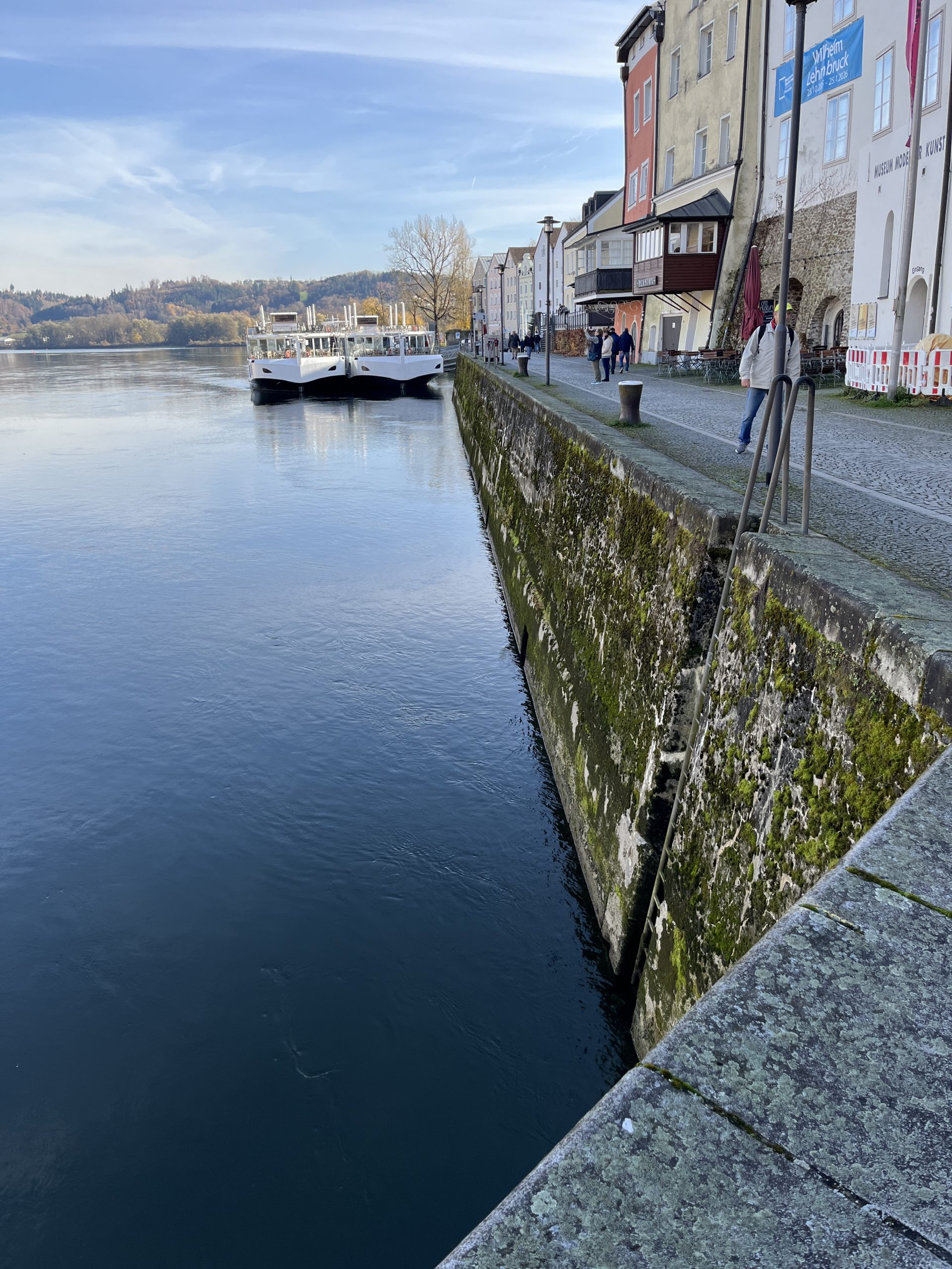 Eine malerische Flussufersiedlung mit historischen Fassaden verschiedener Architekturstile säumt das ruhige Wasser neben einem massiven, moosbedeckten Steinwall.