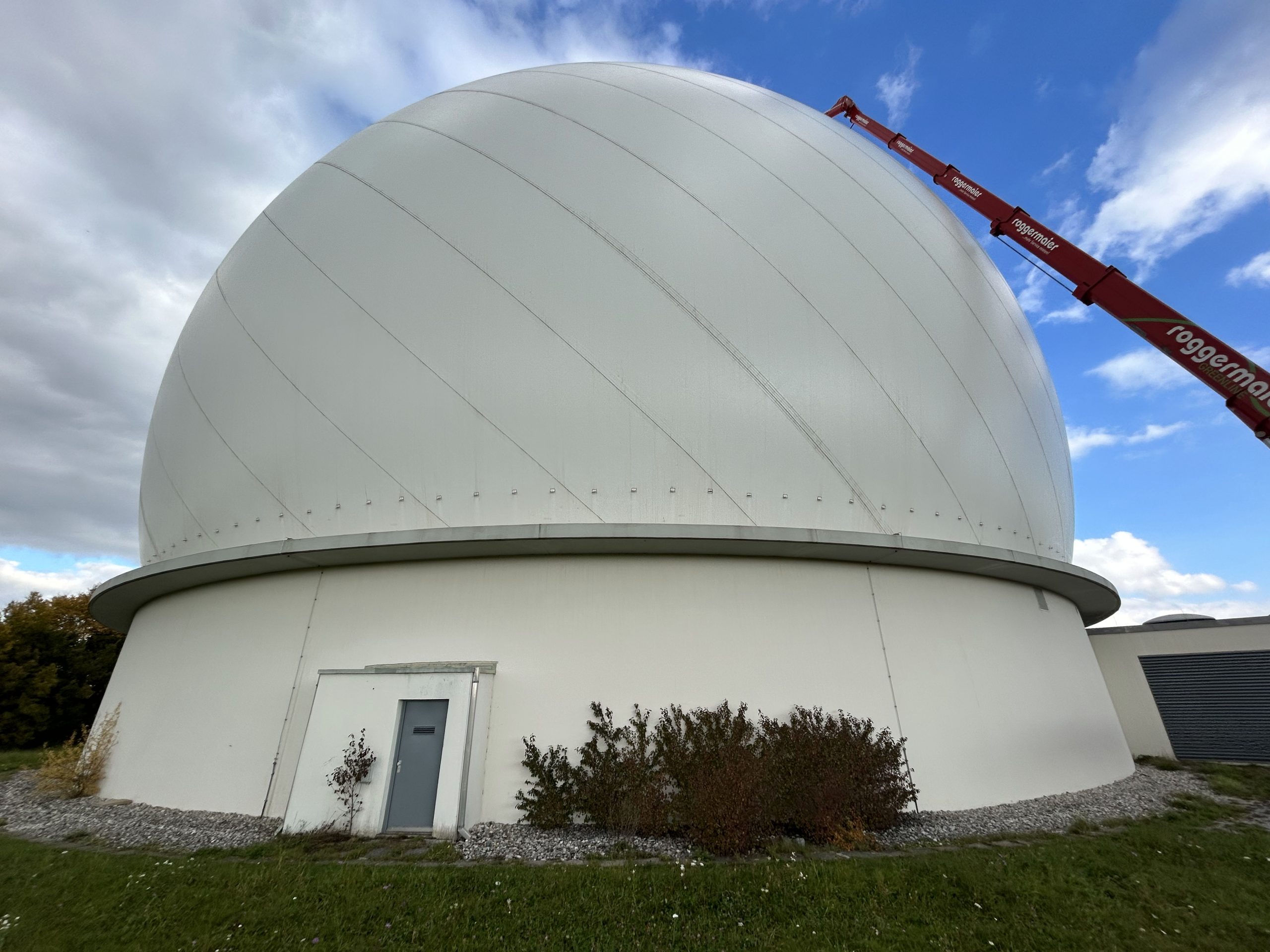 Eine weiße, kuppelförmige Observatoriumskuppel mit horizontalem Äquator-Ring und offenender Luke, davor ein moderner, zylindrischer Betonbau.