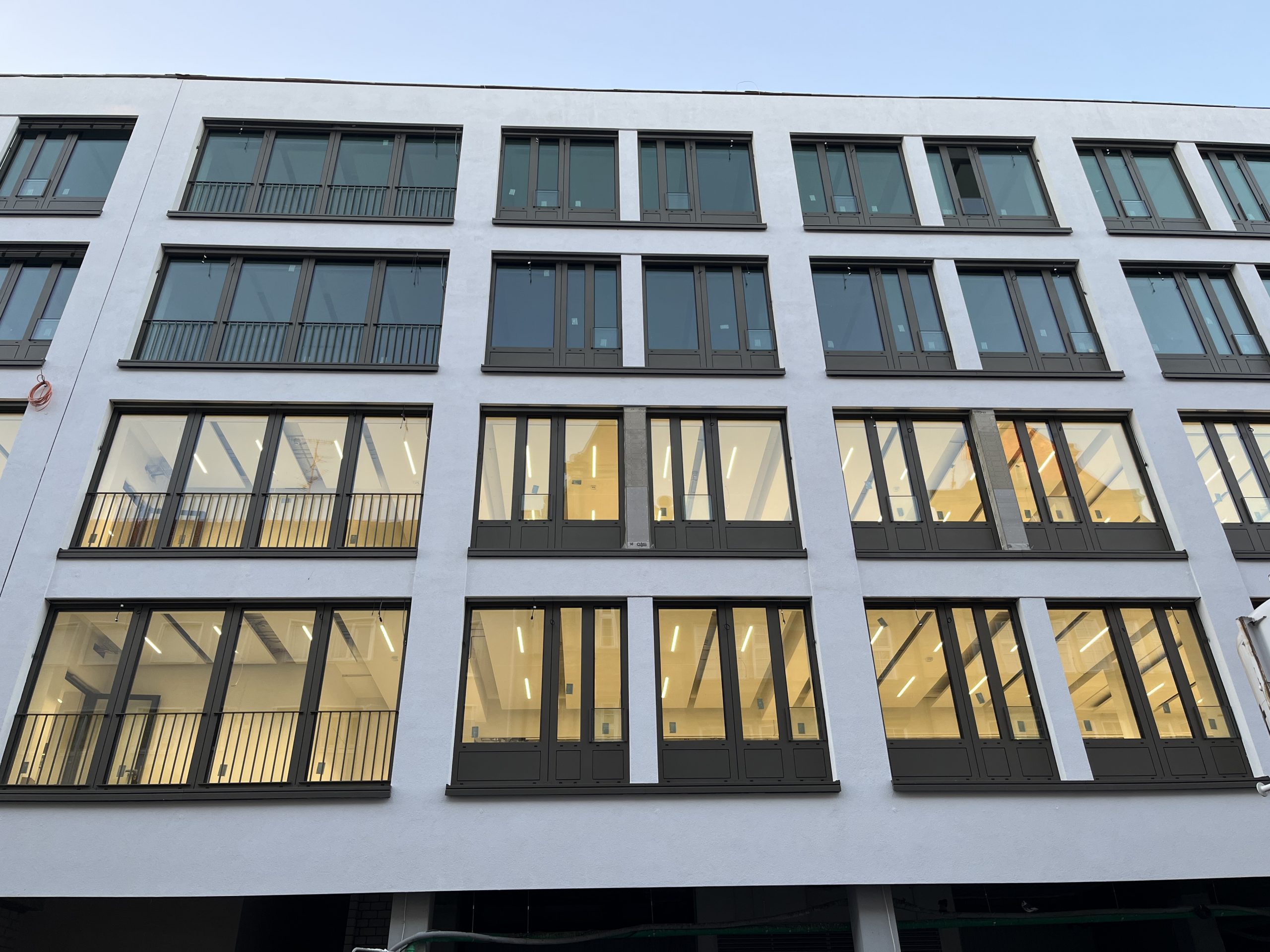 Ein modernes Wohngebäude mit weißer Fassade zeigt regelmäßig angeordnete Fenster mit dunkelgrauen Rahmen und Balkonen.