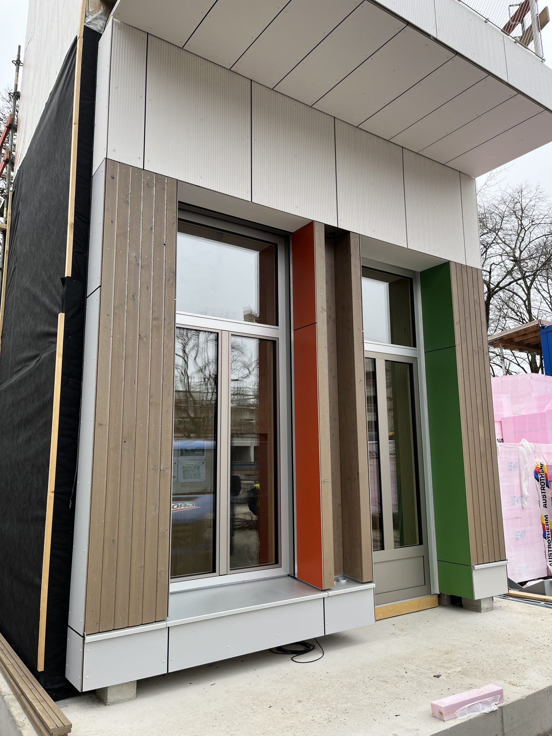 Modernes Modulgebäude mit weißer Wellblechfassade, vertikalem Holzverkleidung in Grau, roten und grünen Akzenten sowie großen Fensteröffnungen.