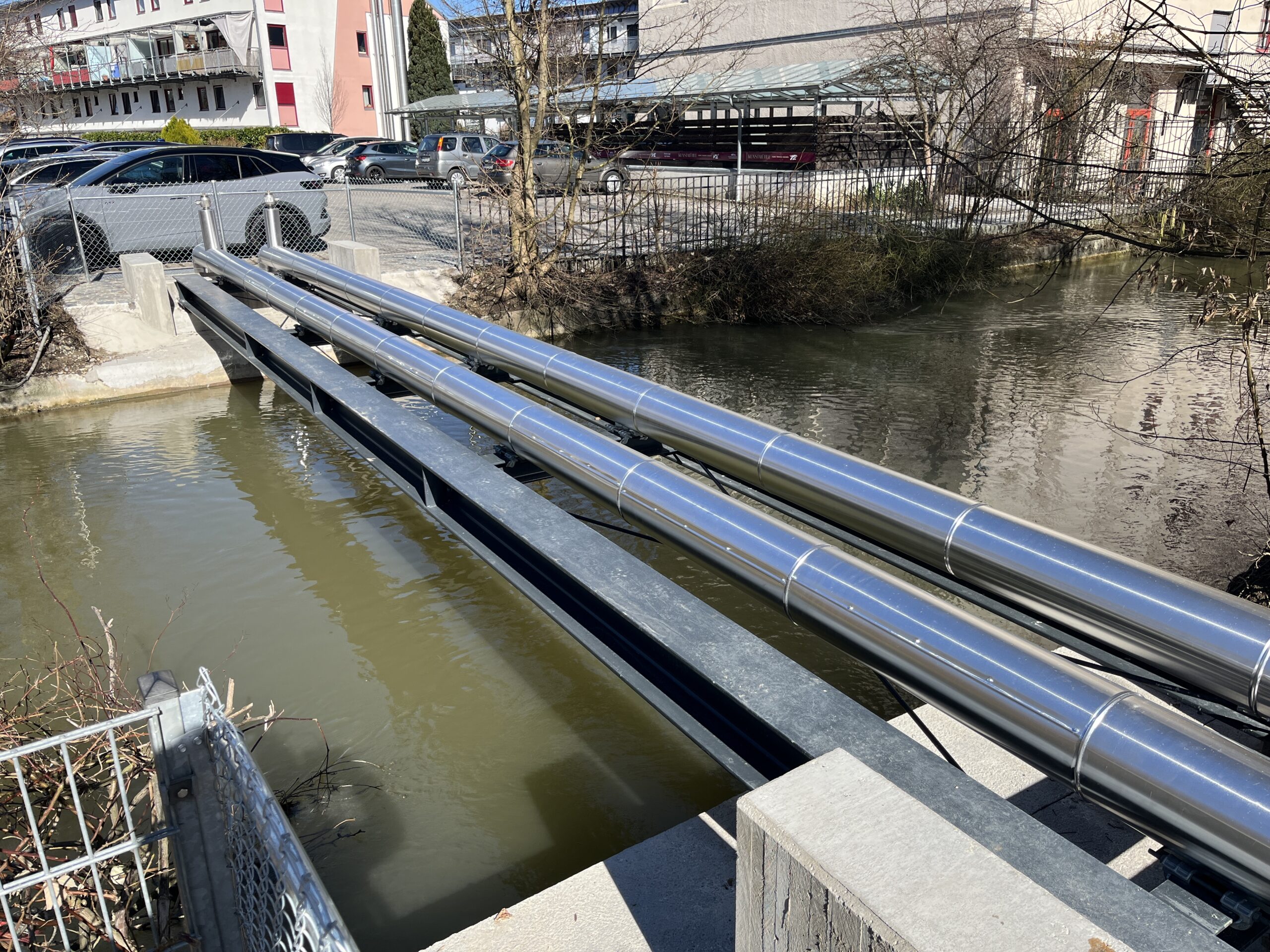 Urbane Infrastruktur: Große blaue Rohre über einem Kanal, dahinter moderne Wohngebäude mit bunten Fassaden und Parkplätze.