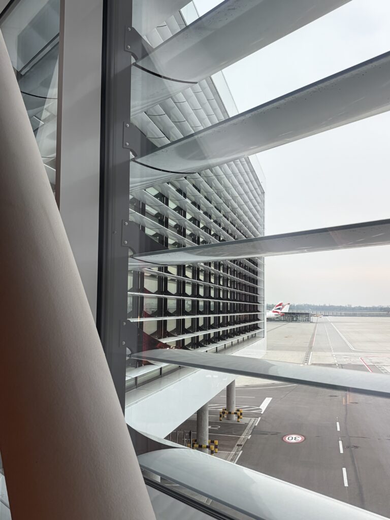 Blick aus dem Flughafen-Terminal durch moderne Glas- und Metallarchitektur auf das ums Eck laufende Terminal und das Vorfeld.