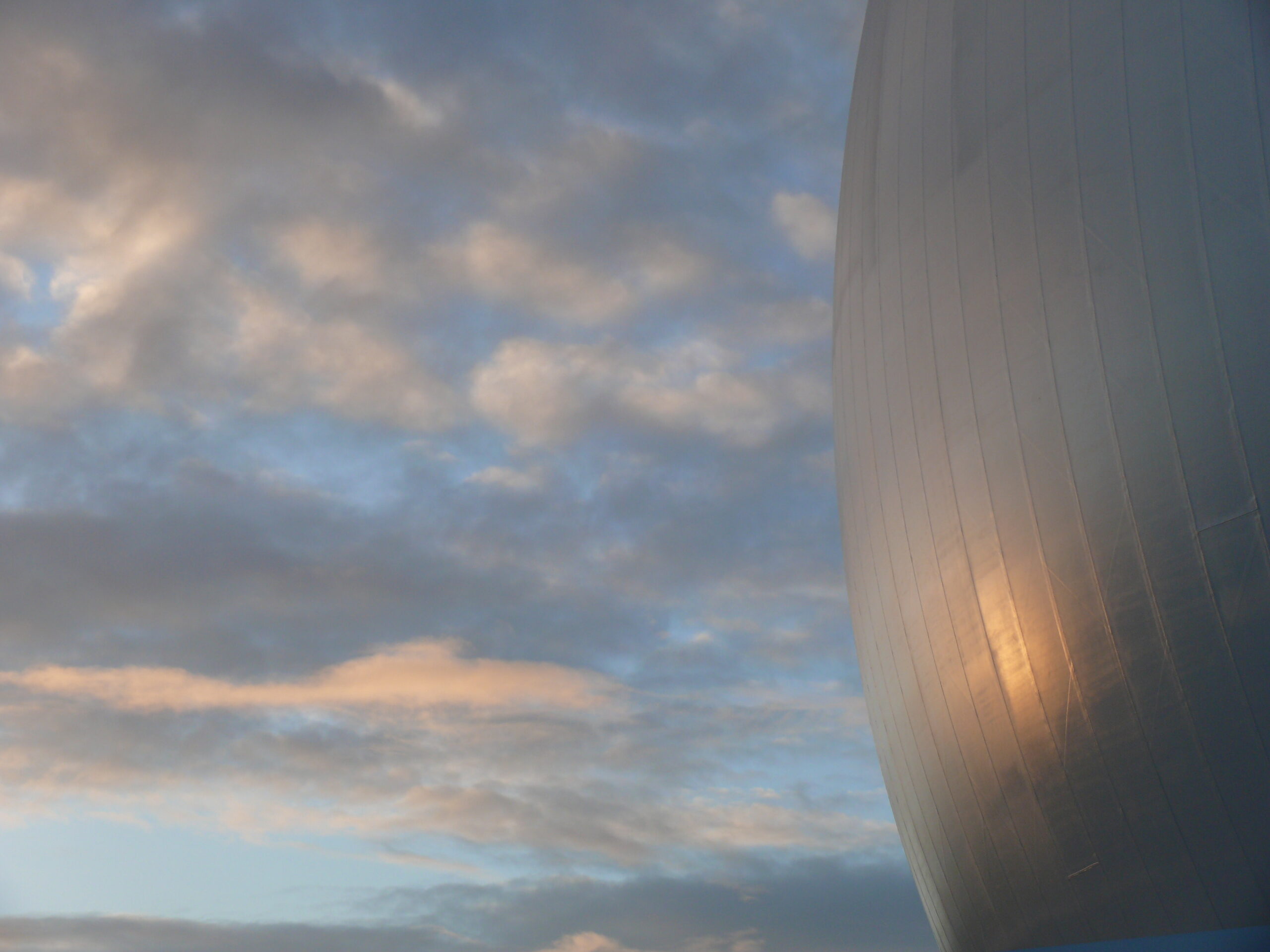 Moderne gekrümmte Metallfassade eines Gebäudes vor bewölktem Himmel. Die silberne Oberfläche zeigt vertikale Rillen und reflektiert das Tageslicht.