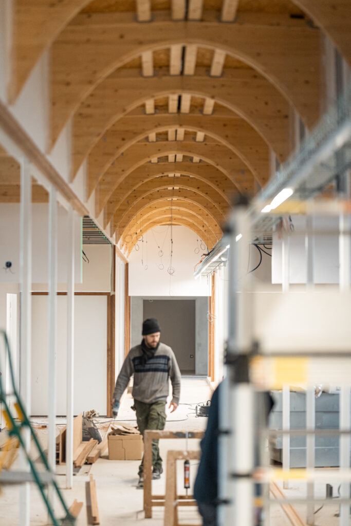 Innenraum im Bau mit sichtbarer Holzdeckenkonstruktion aus gebogenen Trägern, ein Arbeiter bewegt sich durch den unfertigen Flur mit Baumaterialien und Installationen